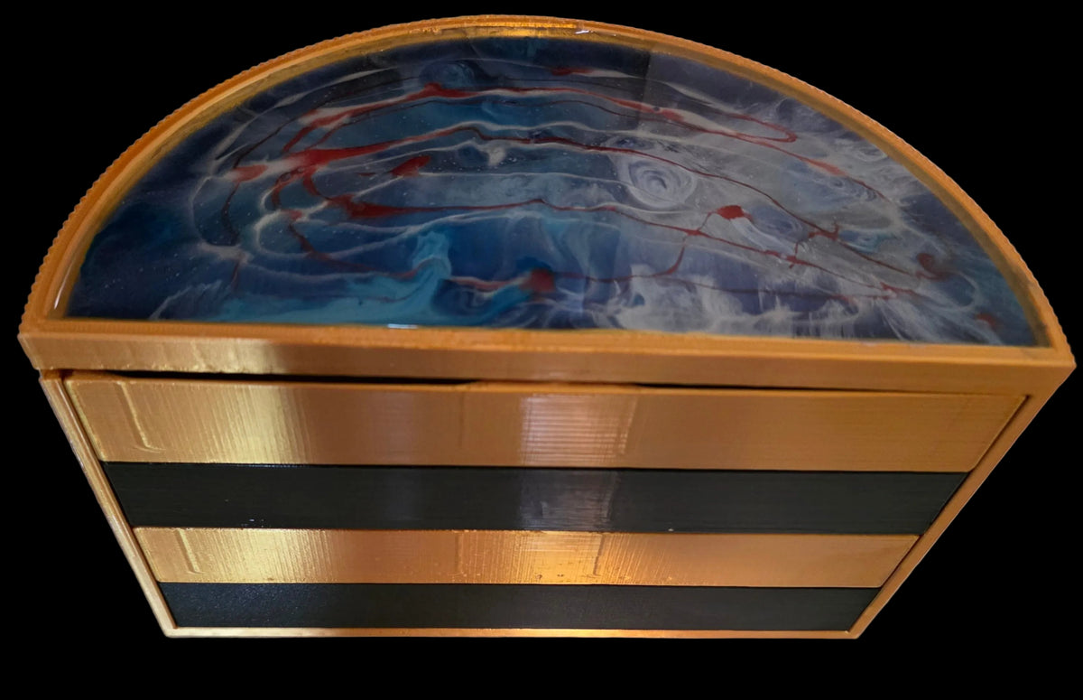 A rectangular storage box with a domed lid featuring a blue, white, and red marbled resin design, framed in gold-toned metal. The front of the box has three horizontal black panels with gold-toned metallic borders.